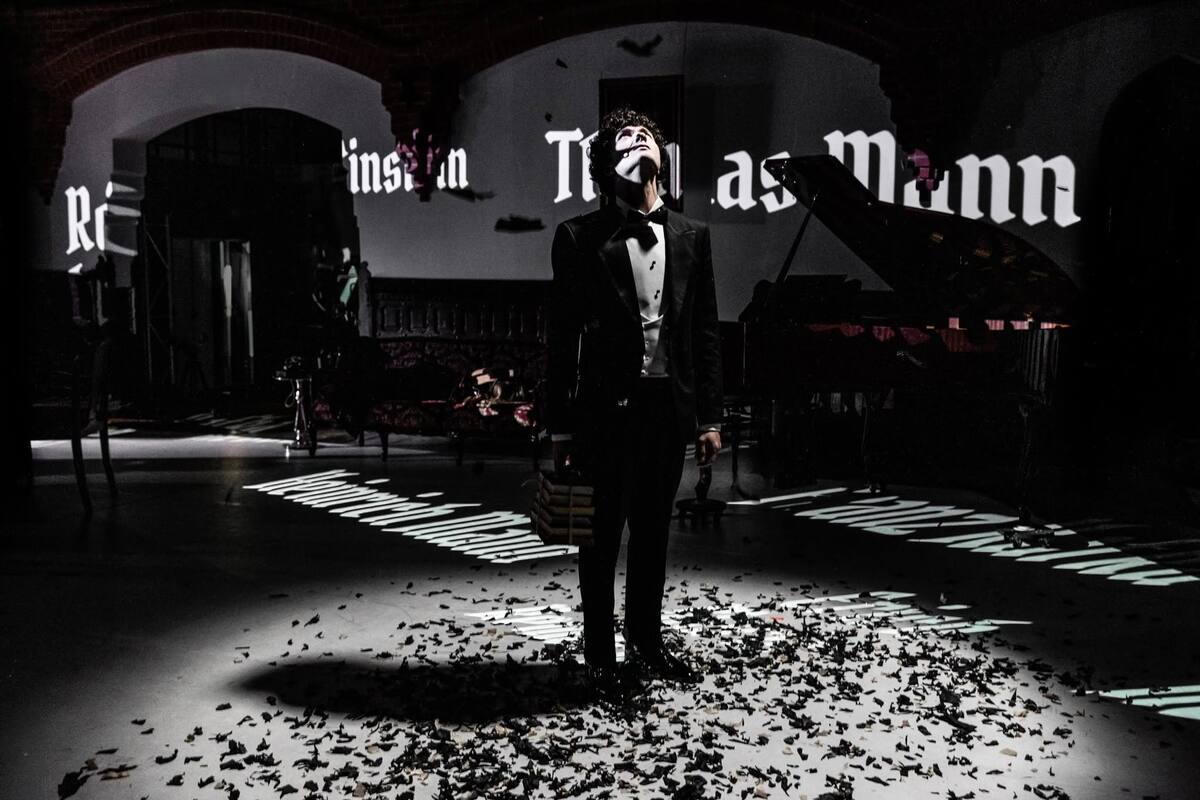 The Damned — a single figure in a beam of light on an ash-strewn floor, names projected across the walls